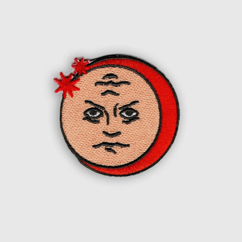 round embroidered patch of a full moon with peach beige background and red crescent with two small red stars. It has a face with angry expression