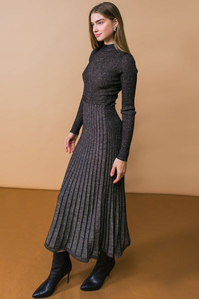 A model wearing a black and silver lurex knit dress with high neckline, long sleeves, fitted waist, and below the knee pleated skirt. Shown from side