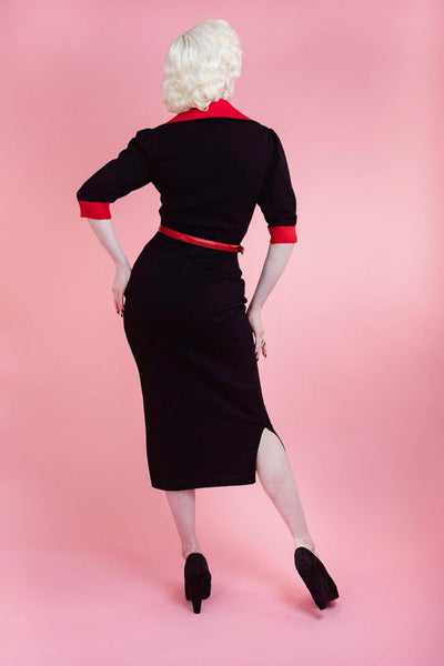 model wearing black sweater knit dress with bright red rolled collar, v-neckline, elbow length sleeves with red cuffs, and embroidered detail across the bodice in matching red of heart-shaped lock, key, and gloved hand. Dress has pencil skirt with side slit and red patent faux leather skinny belt. Shown from back