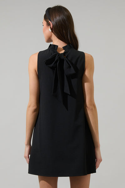 model wearing a black woven sleeveless trapeze dress with high ruffled neckline, pockets, skirt ending above the knee, and self tie at nape of neck. Shown from back