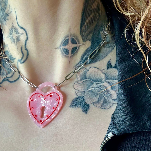 16" silver metal paperclip style chain with pendant in pearly pink marbled acrylic in the shape of a heart-shaped padlock with painted red detail. Shown in close up worn by model wearing leather jacket