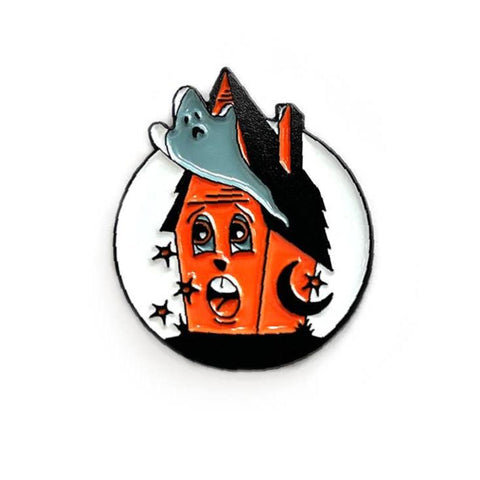 round enamel pin picturing an orange and black haunted house with a scared face & a white and grey ghost flying in front of it