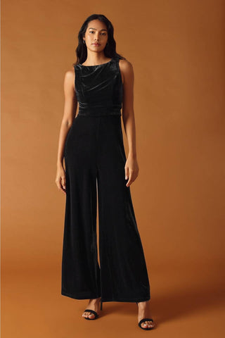 model wearing black velvet jumpsuit with boatneck and seamed sleeveless bodice, banded waistband, and wide legs. Shown from front