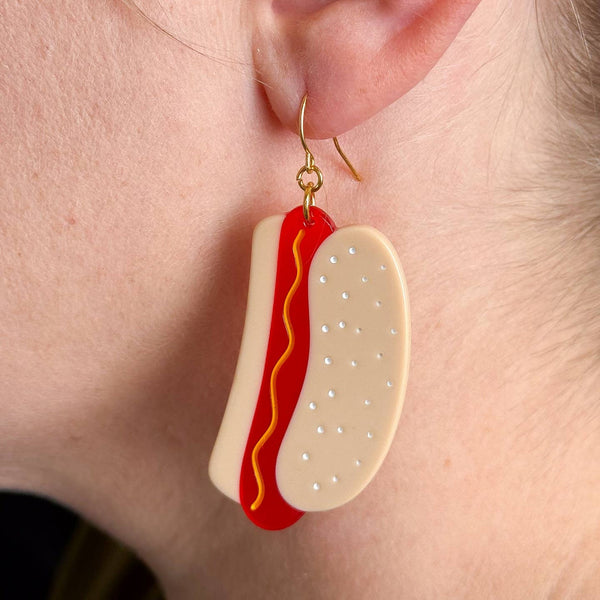 cellulose acetate dangle earrings in the shape of a pair of hot dogs in buns with yellow wavy mustard detail. Shown on model