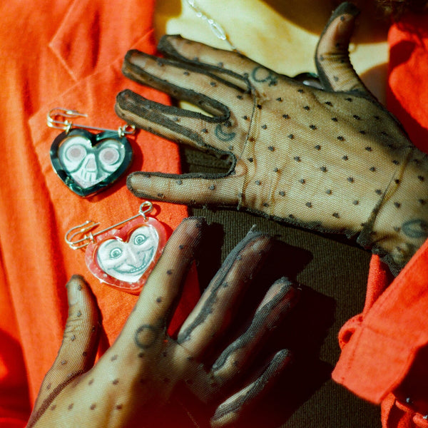 A close-up of a person wearing a bright orange jacket and sheer black polka-dot gloves, holding two heart-shaped safety pin brooches against the chest.
