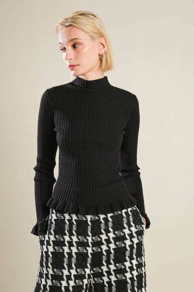 model wearing ribbed knit black mock turtleneck with long sleeves and ruffle detail at each cuff and bottom hem. Shown from the front