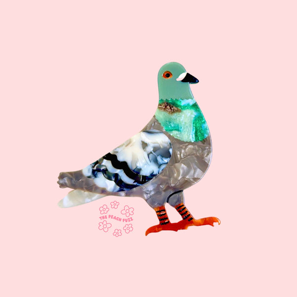 marbled acetate brooch of a pigeon with green head, grey body, and black & white wings with orange legs and feet. Shown from front on pink background