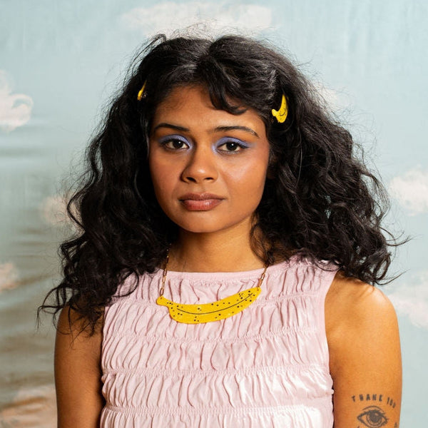 model wearing marbled acetate necklace with large horizontal charm in the shape of a banana with black spots and scattered rhinestone adornment on a gold paperclip style chain.