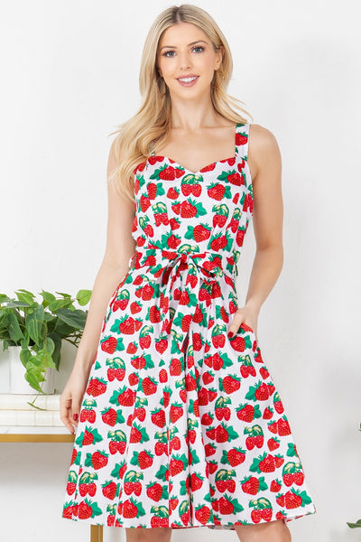 Model wearing sleeveless fit and flare cotton dress with white background all-over pattern of strawberries and raspberries with green leaves. It has a sweetheart neckline, wide straps, princess seaming at the bodice, a self tie at the waist and a flared knee length skirt with pockets. Shown from the front