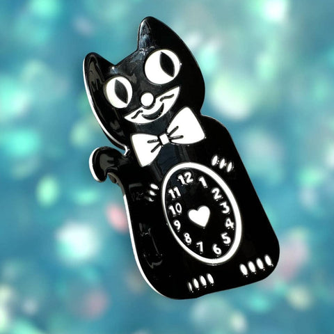 claw style hair clip in the shape of a black Kit-Cat Klock with white details including heart in middle of clock face on its stomach. Shown on glittery blue background