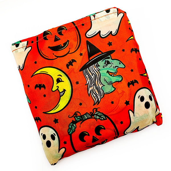 An orange reusable nylon tote bag with an all-over pattern of cartoonish retro Halloween witches, ghosts, crescent moons, and jack-o'-lanterns shown folded in its attached pouch on white background
