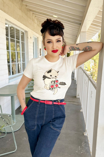 Fitted off-white t-shirt with printed image of a cream-colored kitten with black bow around its neck alongside a blossoming strawberry plant with three berries. Shown worn by a model tucked into denim jeans