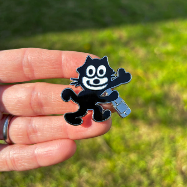 silver metal black and white enamel pin of Felix the Cat smiling and holding a bottle. Shown held against grass background