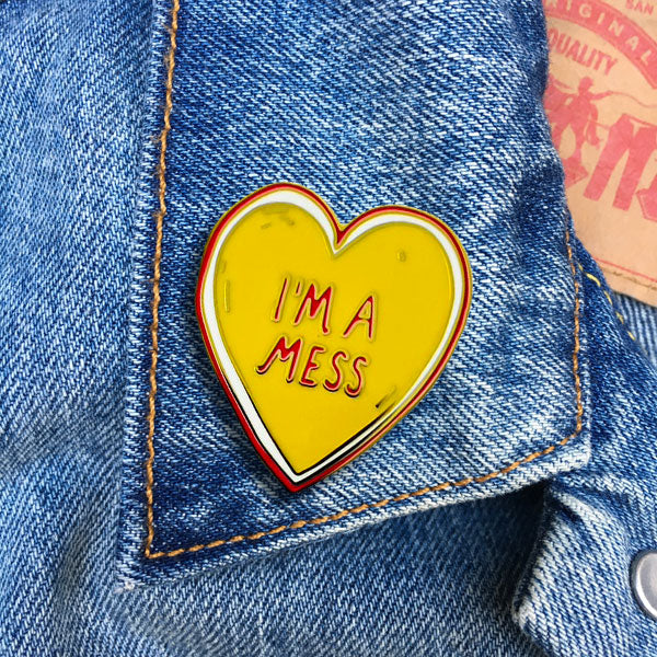 yellow heart with white and red outline and message "I'M A MESS" in red gold metal enamel pin. Shown on denim jacket