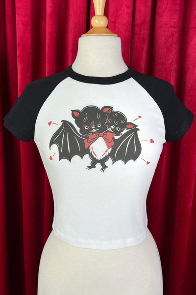 White crop baseball t-shirt with black raglan sleeves featuring graphic of a pair of bats embracing with a red bow on one’s neck surrounded by little red hearts on a mannequin against a red curtain background