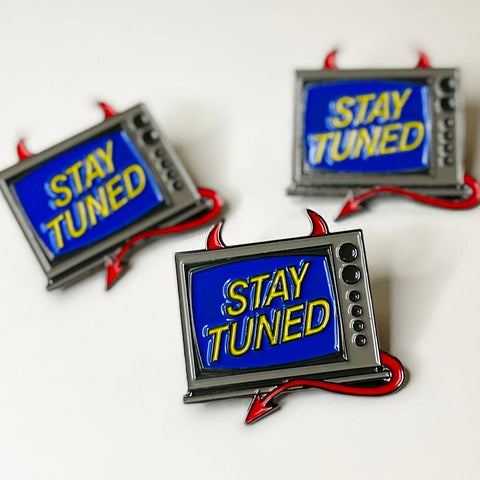 metal enamel pin of television set with red devil horns and tail and blue screen with yellow text "STAY TUNED"