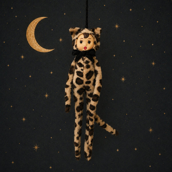 Poseable ornament with wire limbs and Velcro hands in the shape of a pixie with rosy cheeked, green eyed resin face with molded tuft of brown hair. She is wearing a faux fur leopard costume with ears and tail. Black satin ribbon for hanging is at top of head. Shown on black background with gold celestial pattern