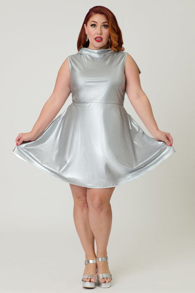Plus size model wearing a metallic silver knit fit and flare mini dress with cowl collar neck with asymmetrical self tie detail, darting at bust, and above the knee length skirt with pockets. Shown from front with skirt held taut
