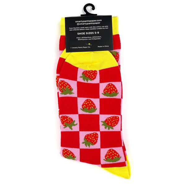 Crew socks with a knit-in pattern of strawberries on a pink and red checkered background with bright yellow cuffs, toes, and heels. Shown flat in packaging from back