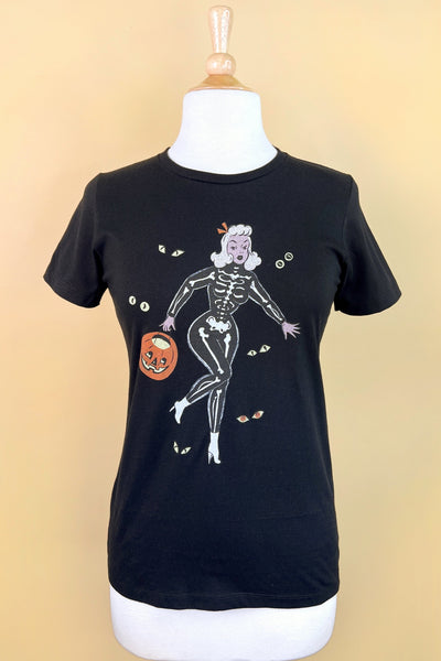 black short sleeve t shirt with a printed illustration of a pinup style woman wearing a skeleton costume holding a pumpkin basket surrounded by pairs of monster eyes. Seen on a dress form 