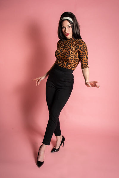 model wearing a pair of black knit cigarette style pants with a high waistband and vertical darting. Shown from the front on a pink background.