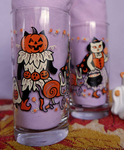 Drinking glasses screen printed with illustrations of cats wearing vintage inspired Halloween costumes. Two shown next to each other on a purple background with one shown in foreground