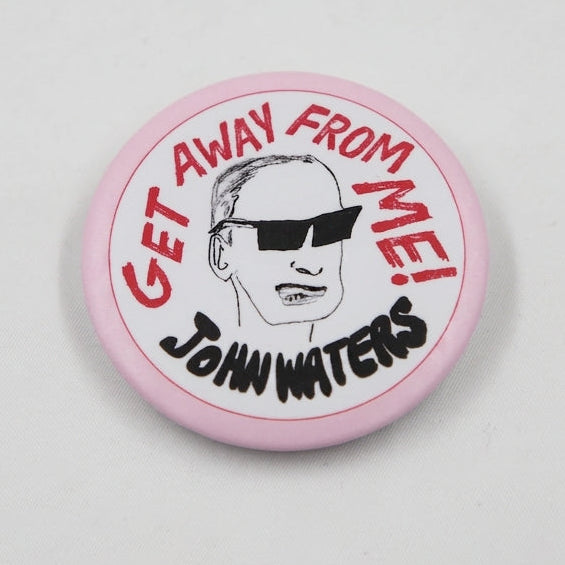 A round pink button badge featuring a black-and-white illustrated portrait of John Waters wearing sunglasses, with red text reading 'GET AWAY FROM ME!' and black text reading 'JOHN WATERS'. Shown from front