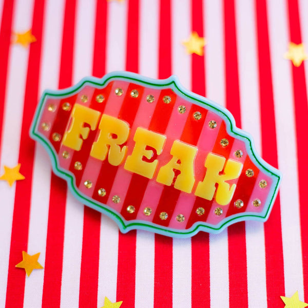 large cellulose acetate barrette in the shape of a circus tent banner with red and pink vertical stripes and blue and green border and "FREAK" in stylized yellow letters surrounded by a row of yellow rhinestones. Shown flat on red and white striped background