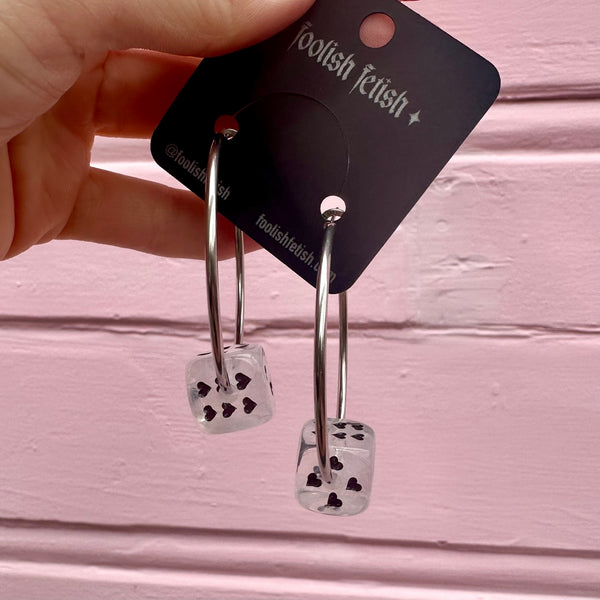 Silver hoop earrings with clear and white marbled dice-shaped charms with black heart-shaped pips on a pink background. Shown from the front