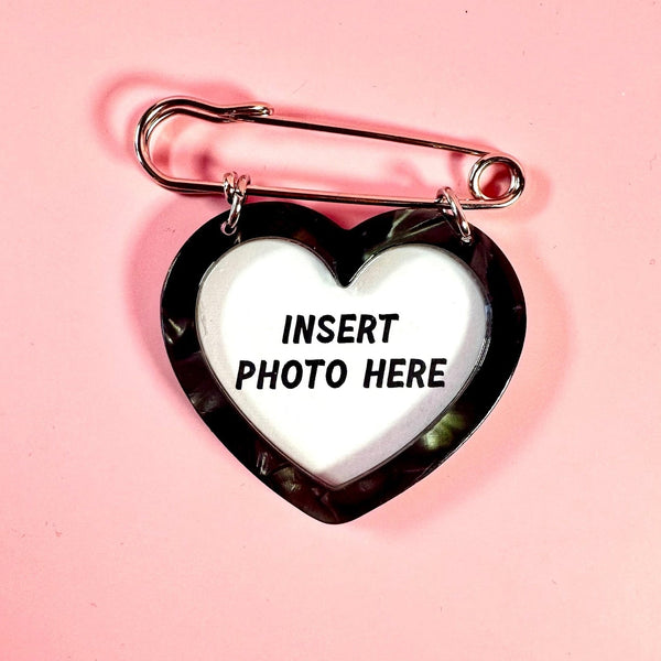 A safety pin brooch with an attached black marbled heart pendant and a white center that reads 'INSERT PHOTO HERE' in black text, photographed on a solid pink background.