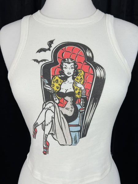 off-white ribbed tank top with round neck and printed illustration of pinup style woman with stitched legs wearing gown and sitting in coffin-style large chair. Printed in red, black, blue, and yellow. Shown on dress form in close up