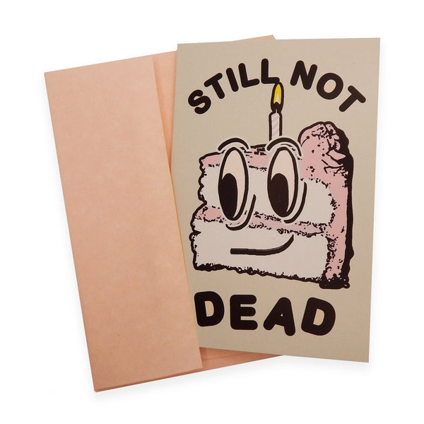 rectangular heavyweight note card with image of smiling cartoon slice of cake with single candle and message "STILL NOT DEAD" in black. Shown with pink envelope