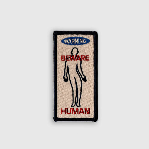 rectangular beige embroidered patch with black border and WARNING BEWARE HUMAN in red and blue with distorted human silhouette in black outline