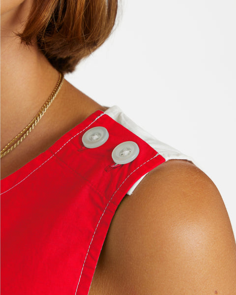 a model wearing a cotton a-line smock style mini dress in half red, half white design divided vertically down the middle of the garment. it has a round neckline with slash detail, button closure running down the left shoulder and down the side of the dress, and patch pockets in alternate colors at each hip. Shown from front in close up of button closure at shoulder