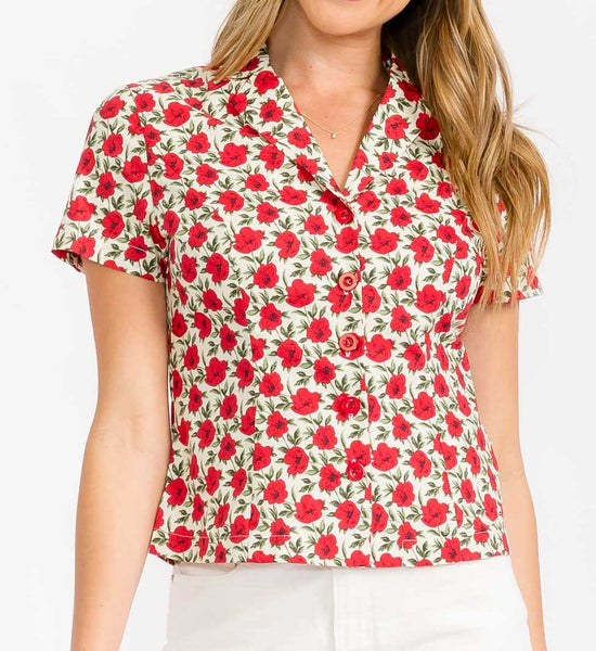 model wearing cotton short sleeve blouse with warm off-white background pattern of red and black flowers with green leaves. Has a notched collar and matching red plastic buttons. Shown from front in close up of bodice