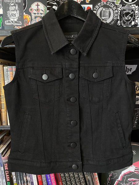 Black denim vest with chest, side, and interior pockets and matte black buttons on a hanger against a background of books and posters. Shown from front