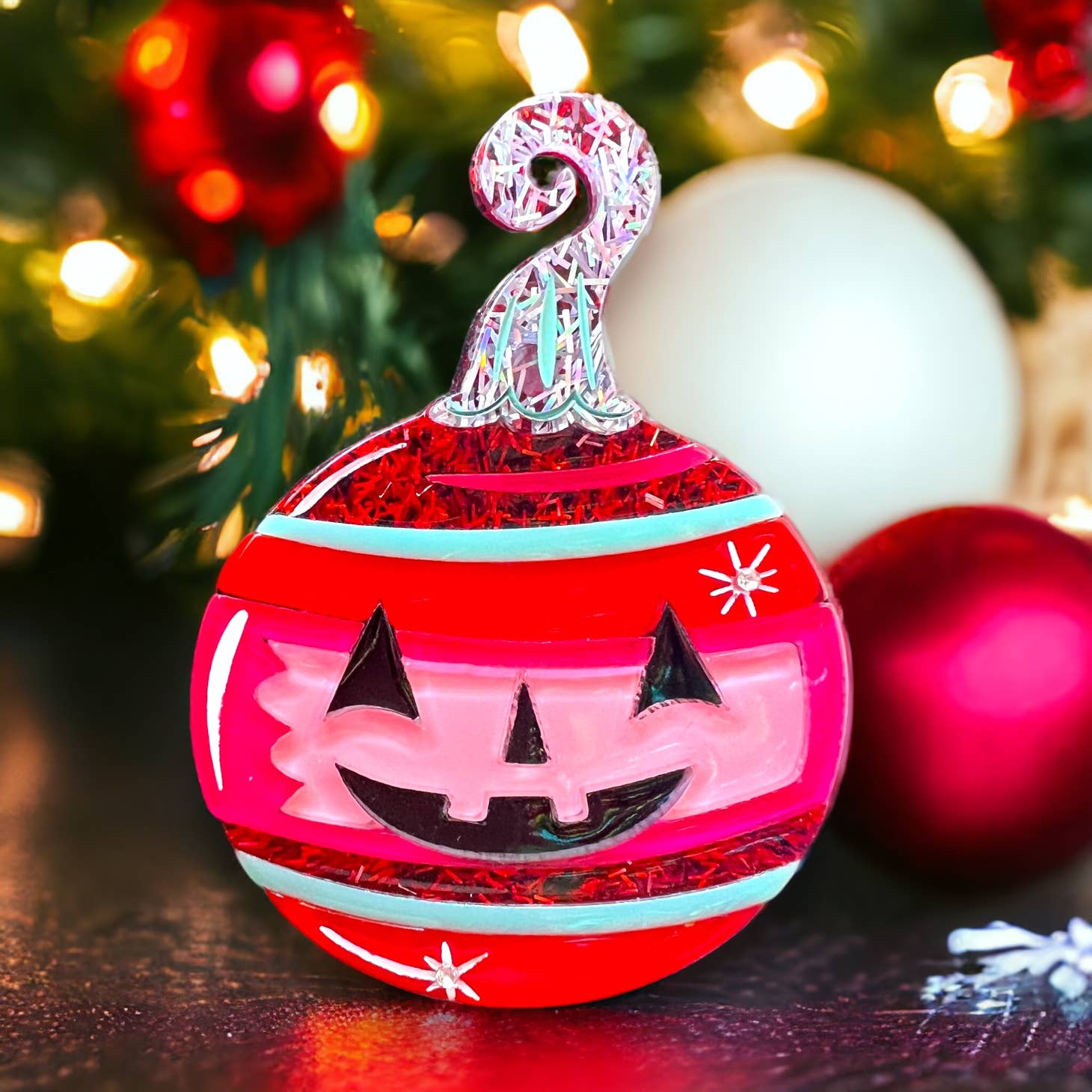 Spooktacular Sparkle Brooch by Johanna Parker x Lipstick & Chrome, tree ornament shaped brooch with black jack-o’-lantern face and green/red/pink stripes with red and iridescent glitter. Shown with Christmas tree in background