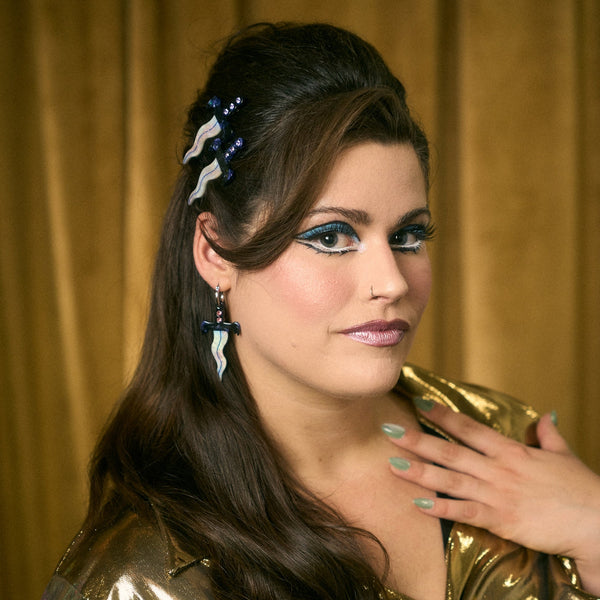 pair of small stainless steel hoop earrings with small acetate dagger charms with wavy iridescent blades and marbled blue and white handles with a row of three rhinestones in each. Shown on woman wearing gold blouse with matching hair clips