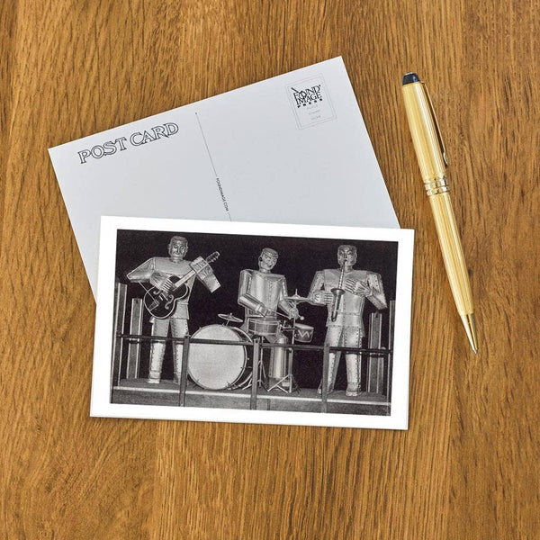 rectangular postcard with black and grey illustration of three robots playing a guitar, drums, and saxophone on a wooden surface with a pen.
