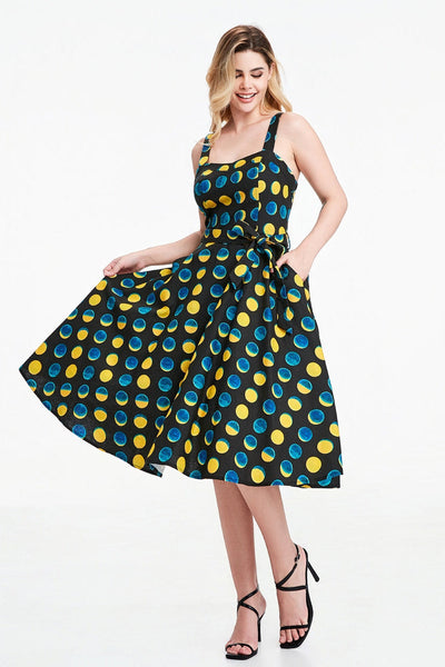A model wearing a sleeveless black background yellow and blue moon phase print dress with a fitted sweetheart neckline princess seamed bodice with adjustable straps, removable self sash belt, swingy gathered full circle just below the knee length skirt. Shown from front with skirt held taut