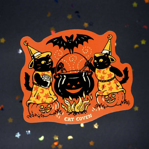die-cut vinyl sticker with orange background of pair of black cats wearing yellow and orange starry witch costumes standing on a pair of jack-o'-lanterns stirring a large pumpkin shaped cauldron being held up by a bat