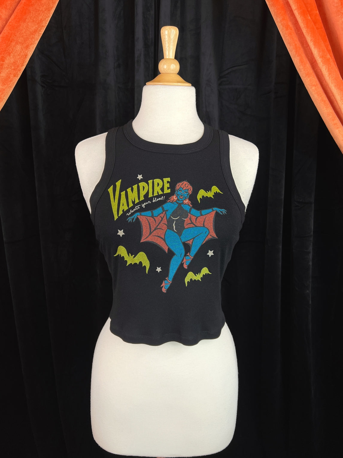 Cropped ribbed tank top with printed illustration of vampire woman with blue skin, red mullet, hoop earrings, a black bodysuit and red spiderweb patterned cape. Message written in yellow and white is "VAMPIRE wants your blood!" alongside yellow bats and white stars. Shown on a dress form