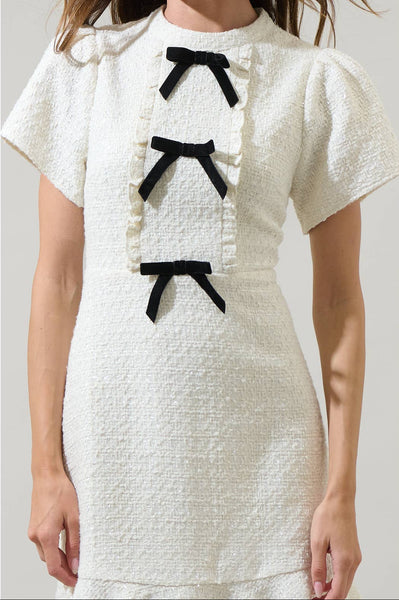 model wearing white textured tweed woven mini dress with mock neck, short sleeves with slightly puffed shoulders, bodice with vertical ruffled bib style and three black bows running down the middle. Dress has a-line skirt ending above the knee in a wide gathered ruffle. Shown from the front in close up of bodice detail