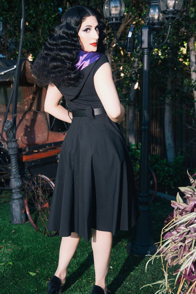 model wearing black bengaline swing dress with cap sleeves, v-neckline, shawl style collar in purple satin and oversized hood lined in matching satin. Dress has black faux leather belt and knee length full skirt. Shown from back to display coffin-shaped belt loops