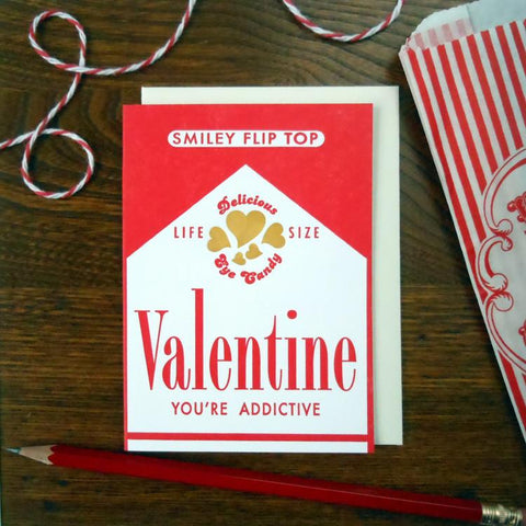 Rectangular letterpress valentines card designed to resemble a package of cigarettes in red, white, and gold. Captioned with “Life Size Delicious Arm Candy” “Valentine you’re addictive”