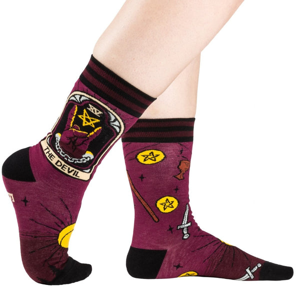 Unisex cotton blend crew socks with striped burgundy and black cuffs and black toes & heels. Each outer ankle has Devil tarot imagery with a hand giving devil fingers alongside a pentagram. Socks have all over pattern of suns, chalices, swords, wands, and pentacles. Shown worn by model