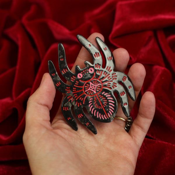 black marbled acetate hair clip in the shape of a spider with red and pink line detail and pink eyes. Shown held in front of red velvet background for scale