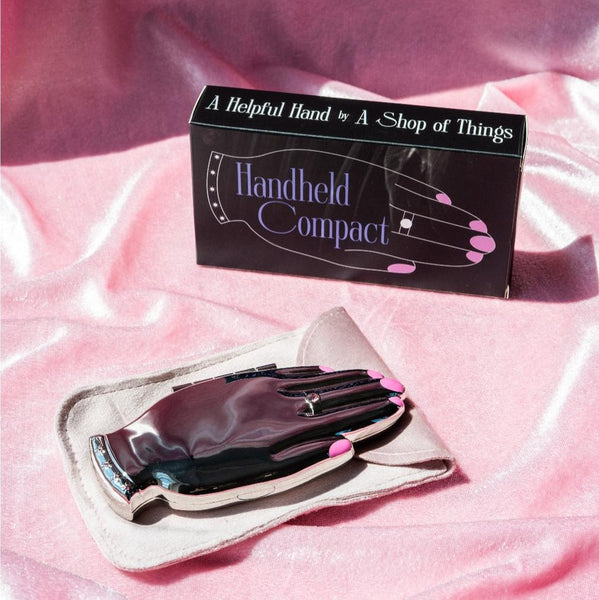 shiny finish silver metal hand-shaped compact mirror with bright pink enameled manicure and pink jeweled ring and bracelet detail. shown closed on pink satin background with gift box and faux suede carrying case
