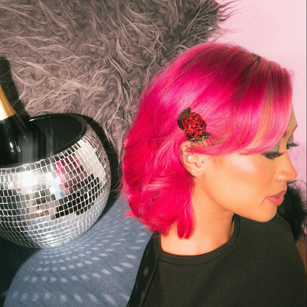 Marbled red and green acetate barrette in the shape of a red rose with green leaves and painted red detail. Shown worn by model alongside disco ball and bottle of champagne