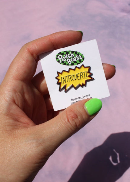 irregular starburst shaped enamel pin with yellow background and red & black outline and word INTROVERT! in black lettering. Shown on backing card